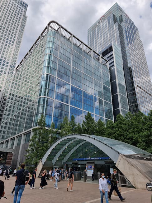 A wide-angle view of the Canary Wharf financial district in London during early evening, showing modern skyscrapers with illuminated windows and distinctive architectural designs. In the foreground, a