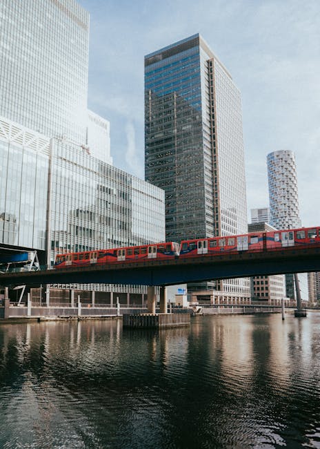 A red train traveling on an elevated railway track crossing over a river in an urban area with modern high-rise buildings in the background. The buildings feature glass facades and varied architectura