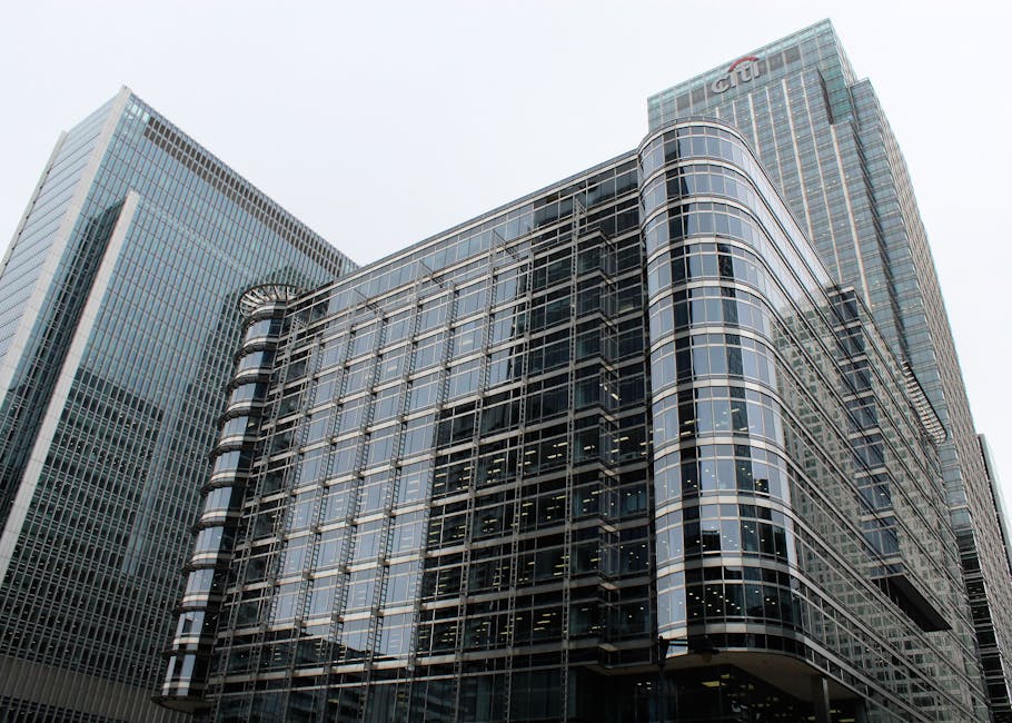 A red train traveling on an elevated railway track crossing over a river in an urban area with modern high-rise buildings in the background. The buildings feature glass facades and varied architectura