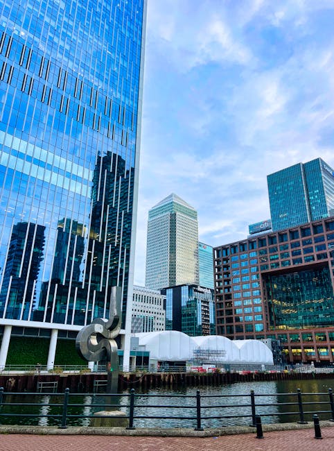 A daytime view of the South Quay area in Canary Wharf showing modern high-rise office buildings and residential towers made of glass and steel, with a clear blue sky overhead. In the foreground, a sec